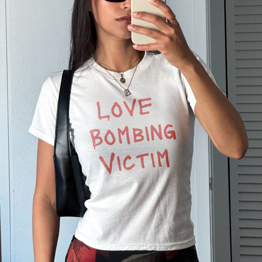 Person wearing a white t-shirt with 'LOVE BOMBING VICTIM' text, taking a selfie.